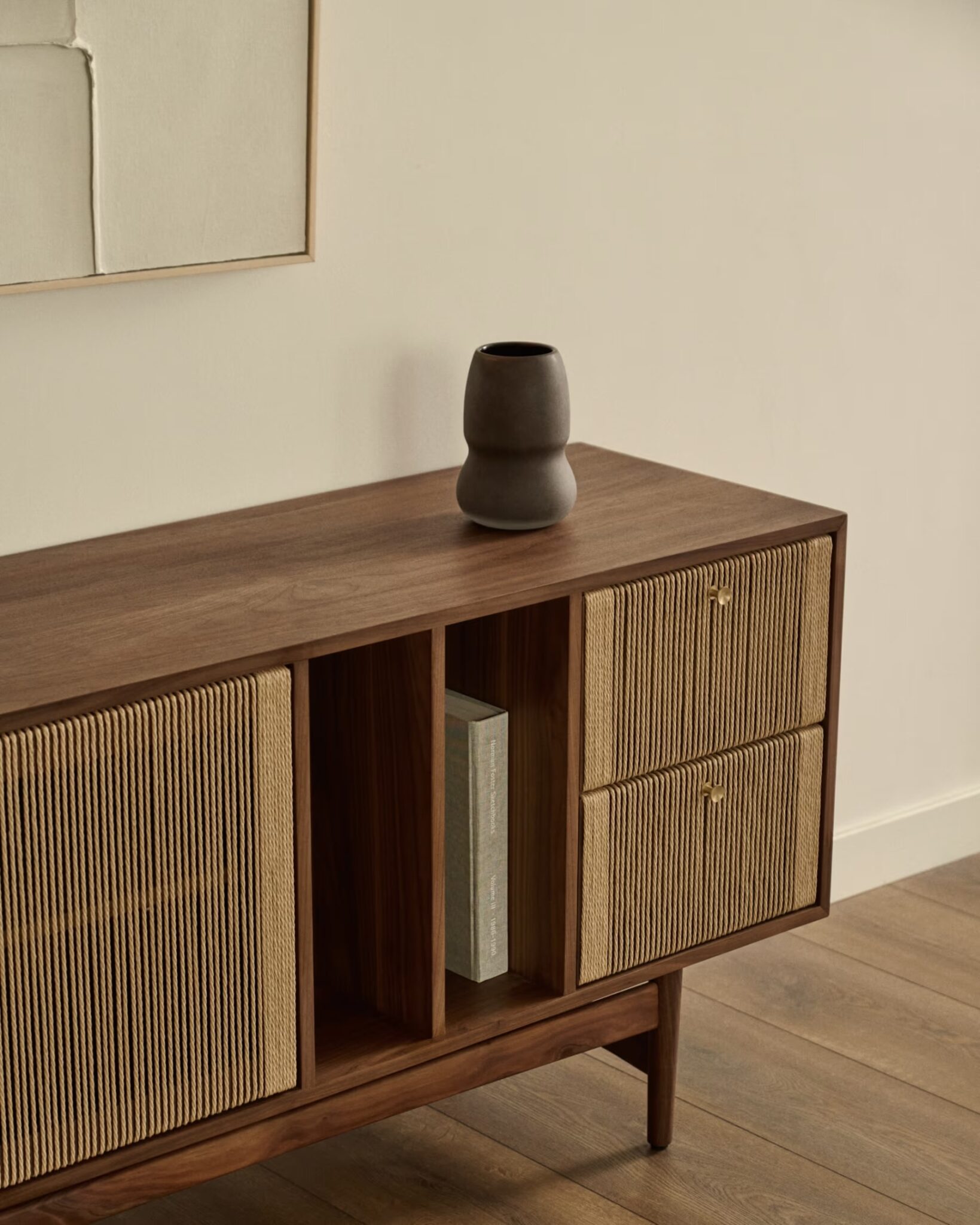 Buffet en noyer avec portes et tiroirs texturés en ficelle, vase gris foncé, livre et art mural minimaliste.